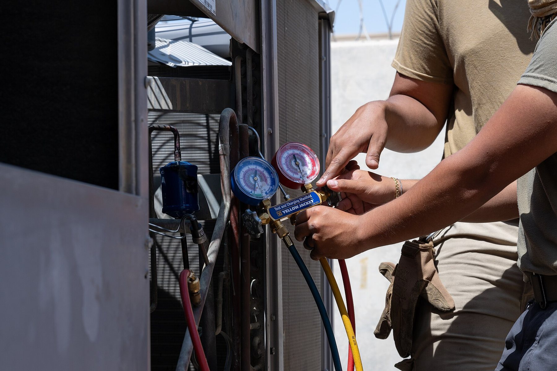 A technician's hands reading manifold pressure gauges on an outdoor condenser unit.
